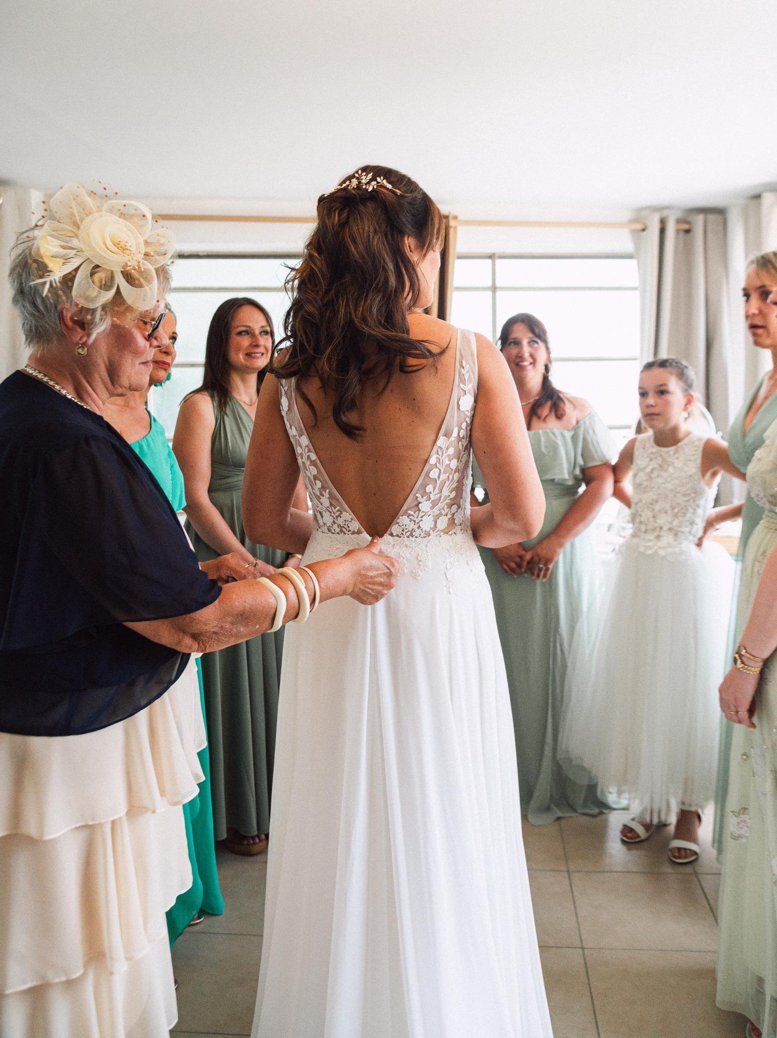 La mariée, vue de dos en robe de dentelle, est entourée de ses proches pendant les préparatifs.